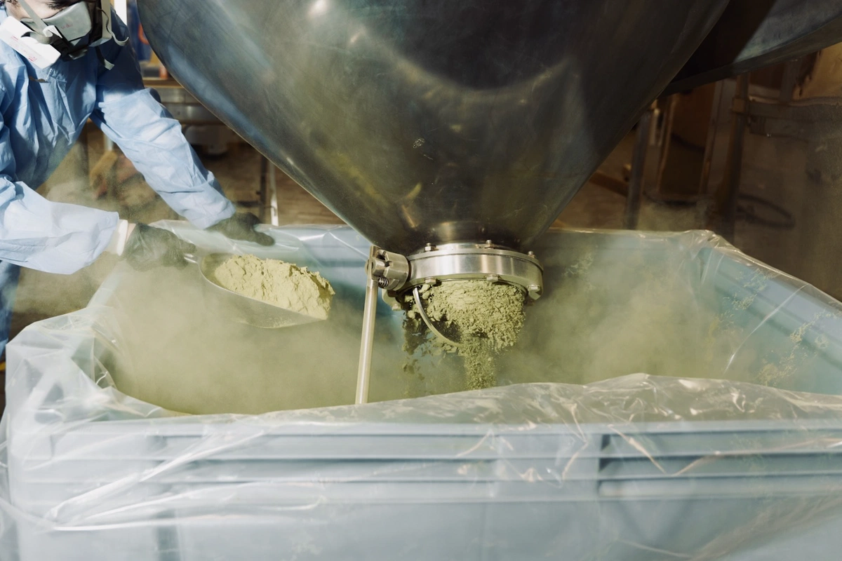 Worker in protective gear filling a large container with kratom powder at an industrial processing facility